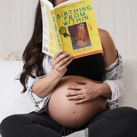 A woman reading a book about birthing with a hand on her pregnant belly